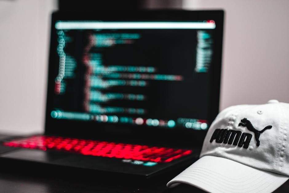 A close-up view of a black laptop with a red-backlit keyboard displaying lines of code on a dark screen, placed on a dark surface. Beside the laptop, there is a white Puma baseball cap with the Puma l